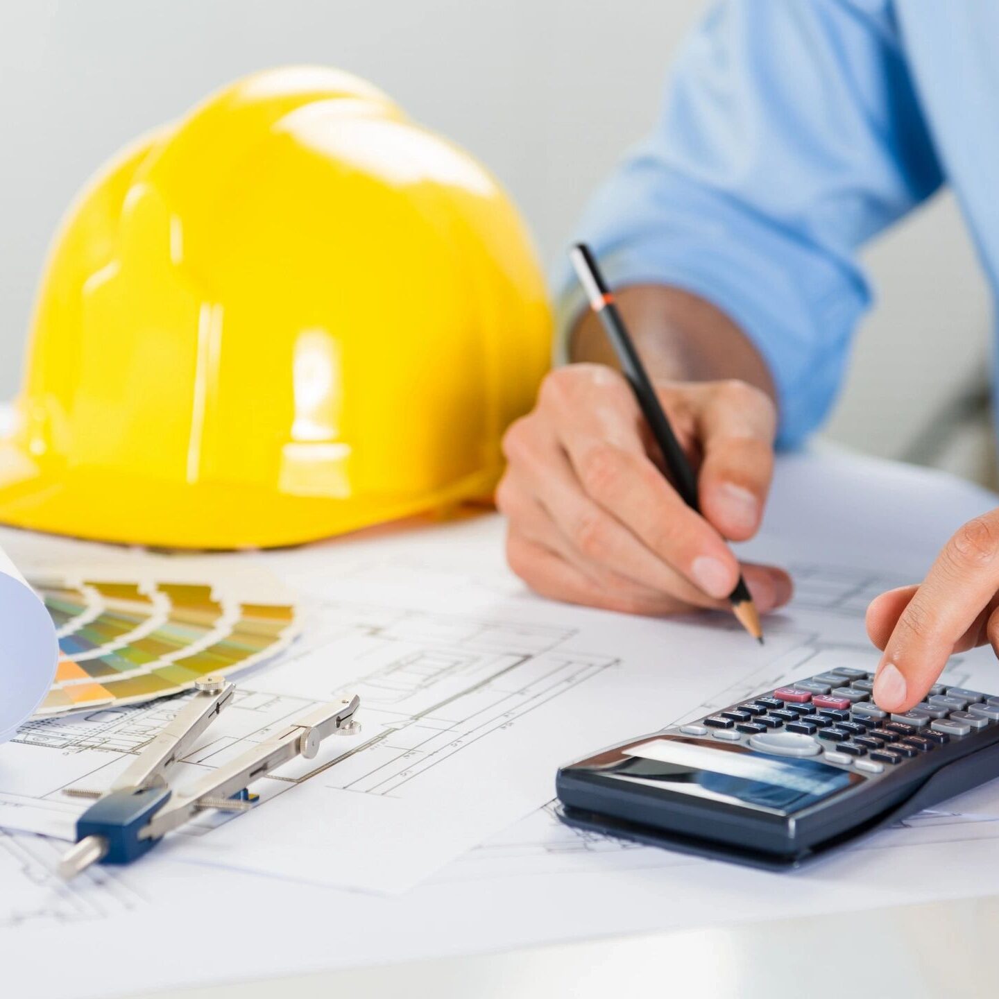 Engineer using calculator and pen over architectural plans with a yellow helmet nearby.